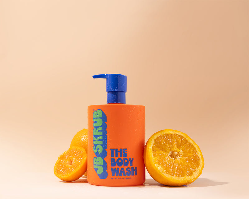 Orange bottle of JB Skrub body wash with blue pump next to sliced oranges on a beige background