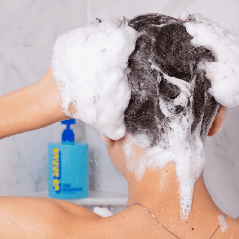 Person washing hair with shampoo, with a bottle of shampoo visible in the background.